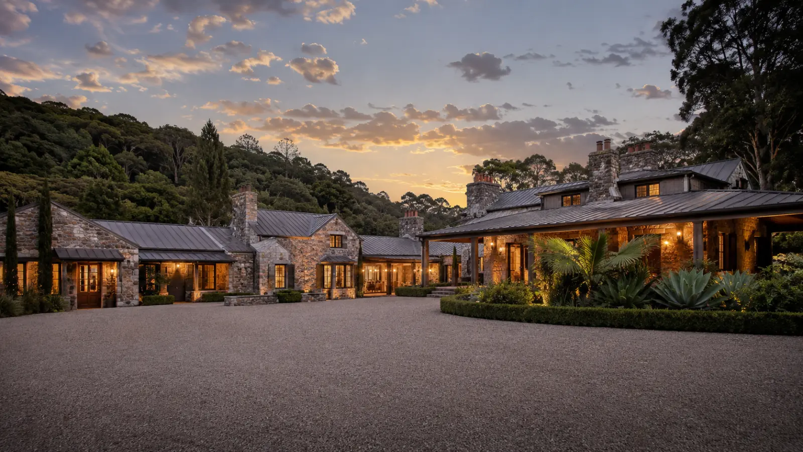 The stone main house at Roy Hinterland lit from within at dusk, above the gravel arrival court